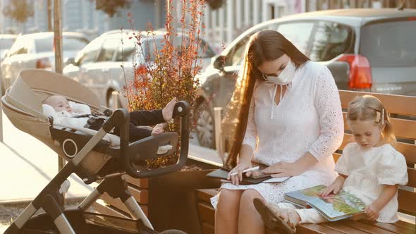 Beautiful Mother in Protective Mask and Two Kids on a Bench in a City