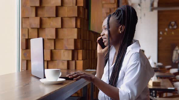 Young Black Woman Talking on the Phone While Sitting in a Cafe with a Laptop