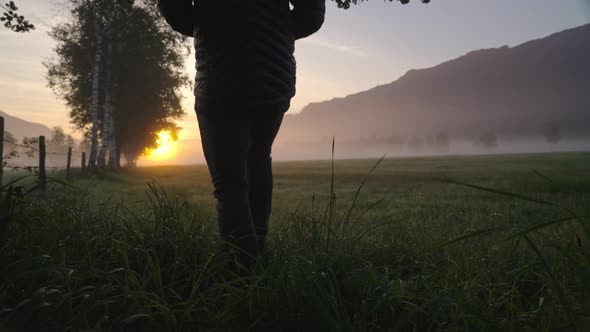 Man Walking Through Meadow Towards Foggy Landscape And Sunrise