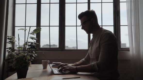 Handsome Cheerful Man with Glasses Typing on Laptop on Against the Big Window