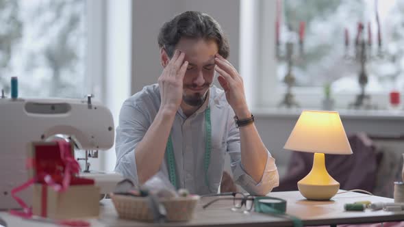 Overworking Male Tailor Taking Off Eyeglasses Rubbing Temples Sitting in Atelier