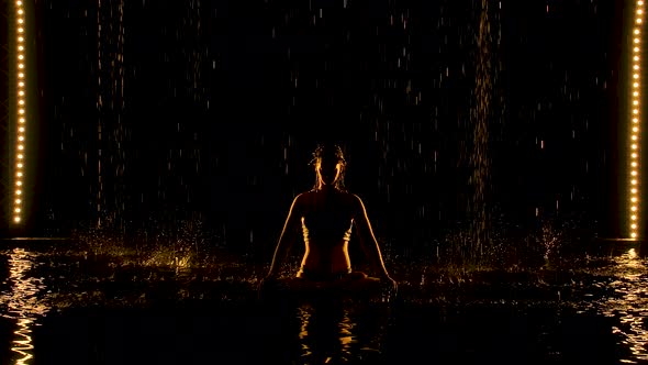 A Young Woman Sits in a Lotus Position in a Dark Studio Under the Rain. A Slim Body Silhouetted with