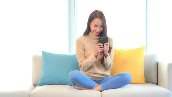 Young asian woman texting or playing video game on smartphone on couch, smiling lady reading news on