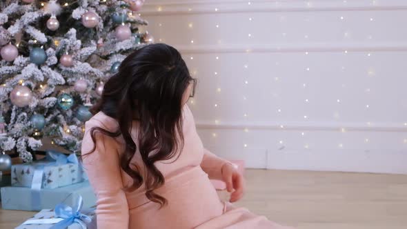 Happy Pregnant Woman Sits Near the Christmas Tree in a Pink Dress and Strokes Her Belly