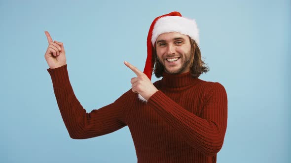 Young Guy in Santa Hat is Pointing Up at Something Winking and Smiling While Posing Against Blue