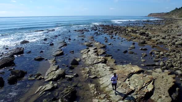 Tracking shot of a young man running on a rocky ocean beach shoreline