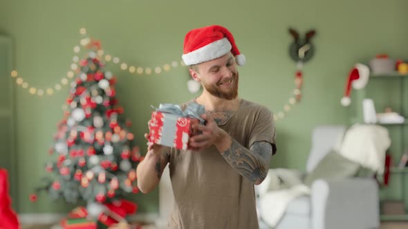 Young Handsome Bearded Tattooed Guy in Green T-shirt with Red Christmas Hat Is Holding a Gift and