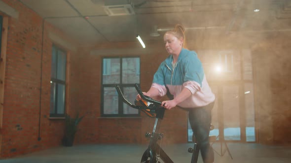 Athletic Girl Performing Aerobic Riding Training Exercises on Cycling Stationary Bike in Foggy Gym