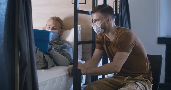 Concentrated Caucasian Men in Covid-19 Face Masks Using Laptop and Talking in Inn or Hostel. Relaxed