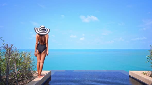 Back View of Lonesome Slender Female in Swimsuit and Floppy Summer Hat Standing by Infinity Pool Wit