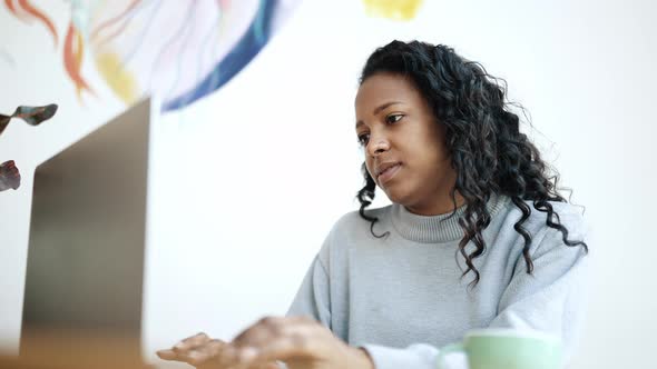 Thinking brunette African woman wearing blue sweater typing on laptop