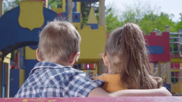 Adorable Blond Boy and a Pretty Girl Sitting on the Bench in Front of the Playground Hugging. A