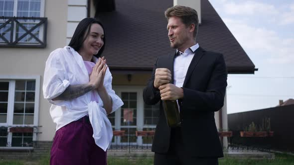 Excited Rich Couple Opening Champagne in Front of New House Outdoors