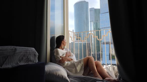 Woman in a Bathrobe Sits on the Floor of a Balcony Against the Backdrop of a Skyscraper During the