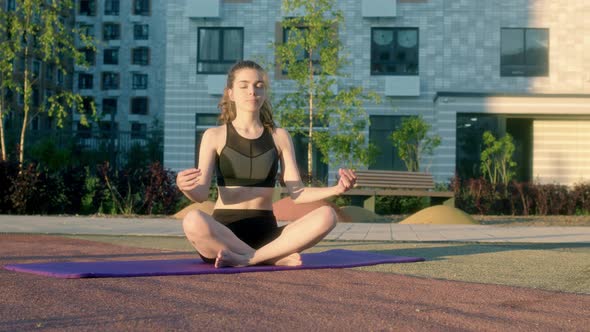 Slender Young Woman in Black Tight Tracksuit Sits on Fitness Mat Outdoors in Residential Complex on