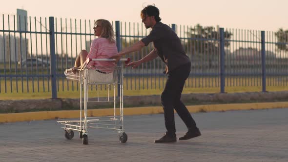 the Guy Rolls Attractive Young Woman in the Trolley on Parking Lot of the Supermarket at Sunset