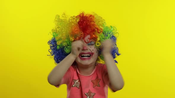 Little Child Girl Clown in Colorful Wig Making Silly Faces, Crying. Halloween. Yellow Background