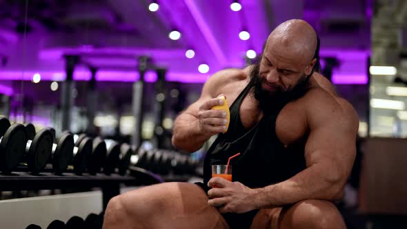 A Brutal Male Bodybuilder Sits in the Gym and Squeezes Fruit Into a Glass with His Hand Drinks Juice