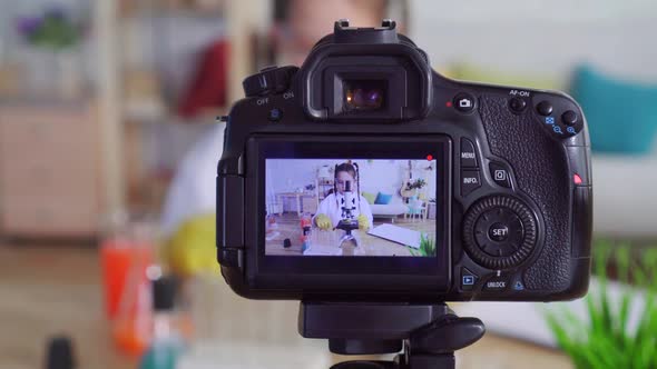 Girl Teen Blogger Youtuber in a White Coat and Goggles in Lab Homework Looking Into a Microscope