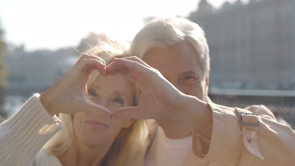 Cheerful Senior Couple Making Heart Shape with Hands Sitting on Bench