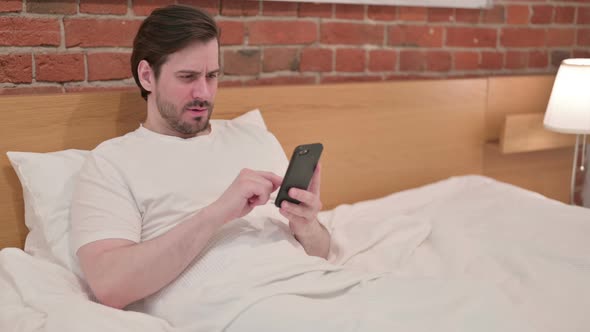 Casual Young Man Reacting to Loss on Smartphone in Bed