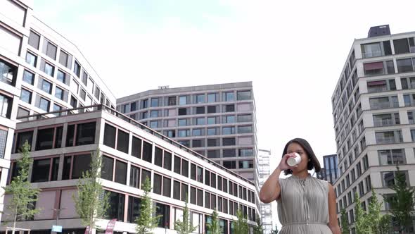 Young business woman drinks coffee to go while walking between tall office buildings in financial di