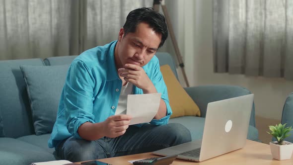 Asian Man Looking At The Bill And Thinking Before Working With A Laptop At Home