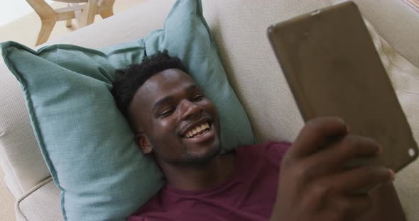 Video of happy african american man lying on sofa and having video call on smartphone