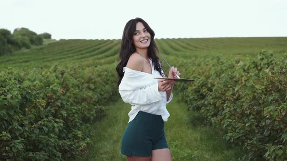 Dazzling Brunette Smiles and Poses with Palette and Brush in Green Field