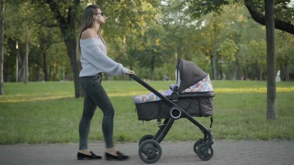 Side View Wide Shot of Gorgeous Confident Woman in Sunglasses Pushing Baby Carriage As Walking in