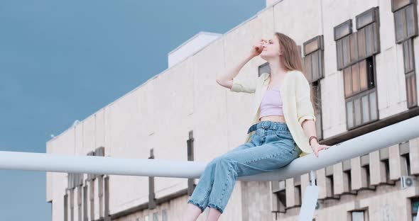 Young Beautiful Woman Sits on Balcony on Buildings Background and the Wind Blows Her Hair