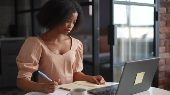 Focused AfricanAmerican Female Student Sitting at Desk Looking at Computer Screen Making Notes