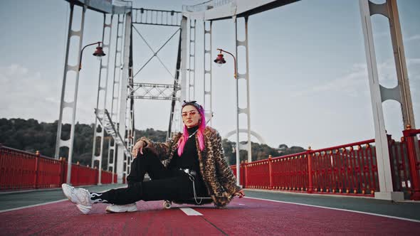 Gorgeous Hipster Woman with Pink Hair in Informal Outfit is Sitting Sideways on Skateboard While