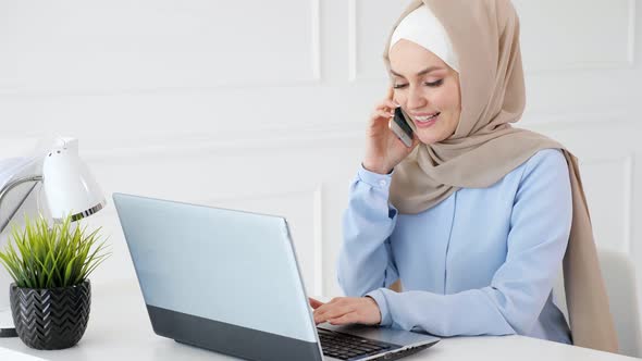 Muslim Woman in Hijab is Talking Mobile Phone and Typing on Laptop at Office Work