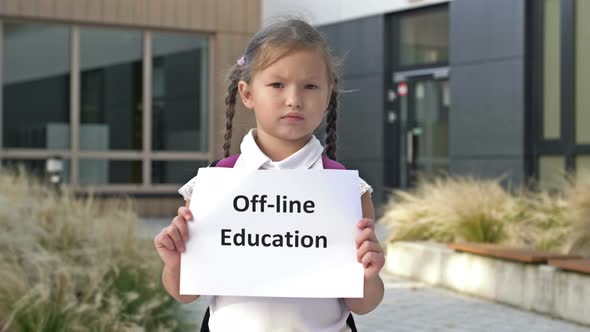 Serious Little Schoolgirl with Pigtails Stands with OFFLINE EDUCATION Poster Outside the School