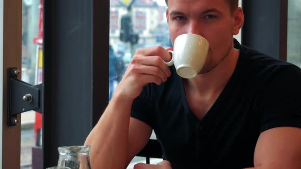 A Young Handsome Man Sits at a Table with Meal in a Cafe, Drinks Tea and Smiles at the Camera