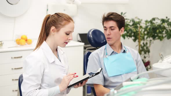 Young Female Dentist Looking at Digital Tablet and Listening Male Patient in Dental Clinic