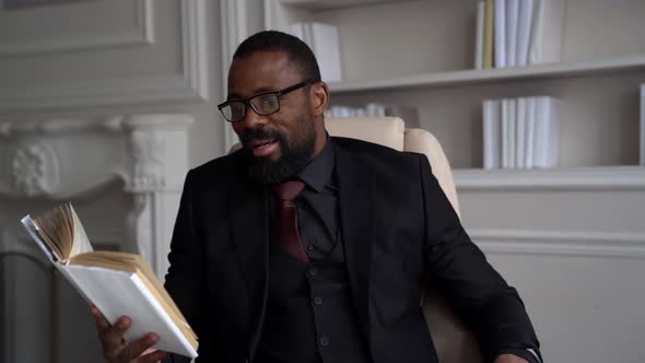a Bearded AfricanAmerican Man in Glasses and a Suit with a Tie is Sitting on a Light Chair in the