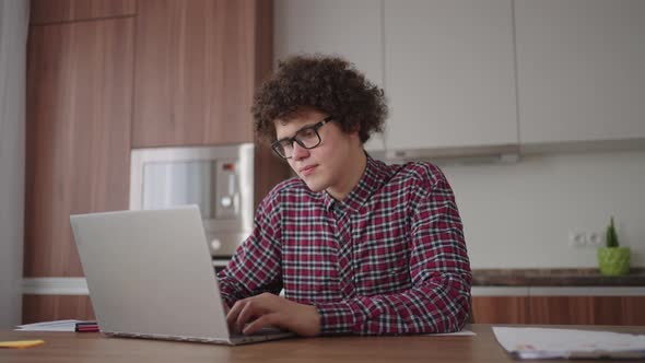 A Curly Man with a Serious Look Works at a Laptop Sitting in a Modern Kitchen