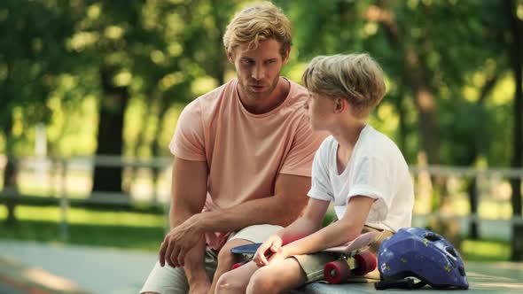 A handsome father is talking to his son after riding a penny board at the park at summer