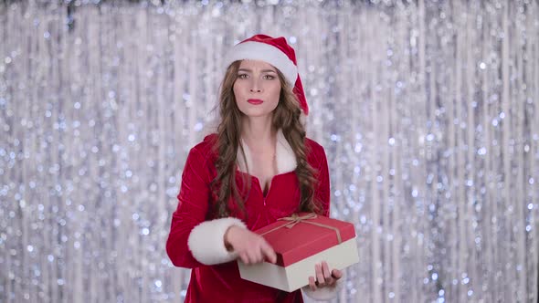 Santa Girl with a Gift in Her Hands Shows Her Finger More Quietly. Bokeh Background