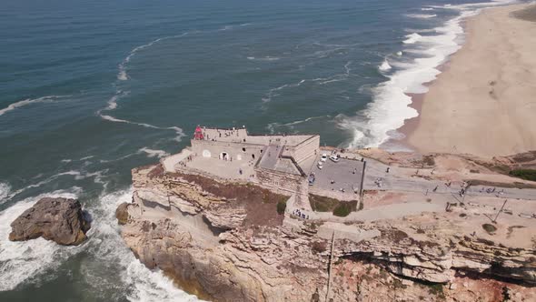 Nazare lighthouse and Saint Michael the Archangel fort perched atop of rocky cliff, aerial pan shot.
