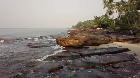 A slow drone shot flying over rocks hitting by upcoming waves from the ocean, Koh Kood Thailand