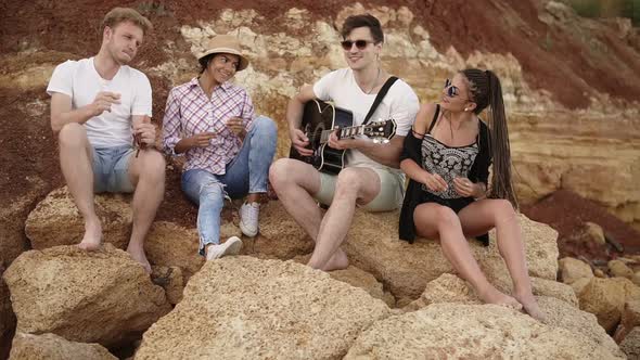 Group of Young Hipster Friends Sitting on the Rocks By the Seashore and Playing Guitar Singing Songs