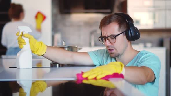 Cheerful Man in Headphones Wiping Cooking Top with Rag and Wife Cleaning Furniture on Background
