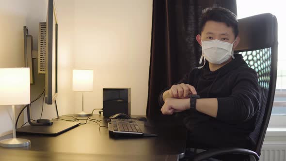 A Young Asian Man in a Face Mask Works on Computer, Then Looks at the Camera and Taps at His Watch