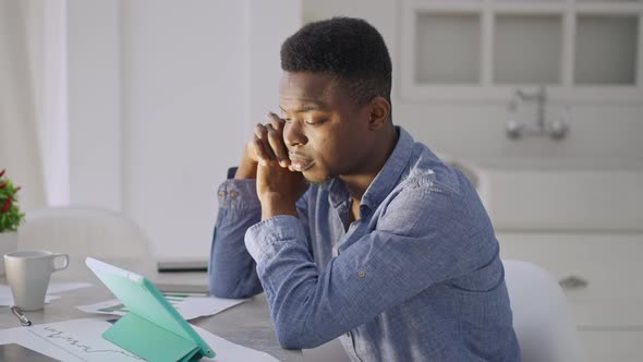 Side View of Tired Young Man Holding Head in Hands Sitting at Table with Tablet in Home Office