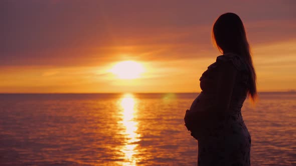 Young Pregnant Woman with a Big Belly Looks at the Majestic Sunset Over the Sea