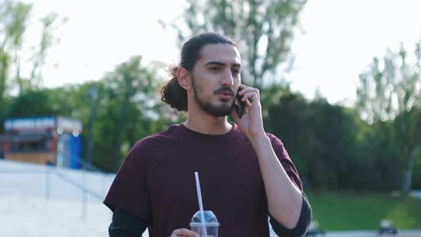 Portrait of Young Attractive Arab Man Talking on the Phone in Park and Drinking Coffee Tonic