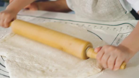 Little Girl Rolls Dough with Wooden Pin at Table in Kitchen